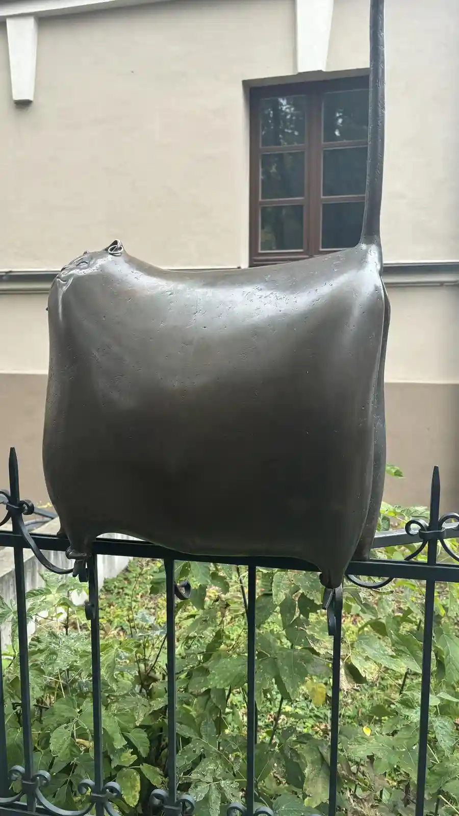 A bronze statue of a cat with an extremely inflated, balloon-like body is perched atop a black metal fence, with green…