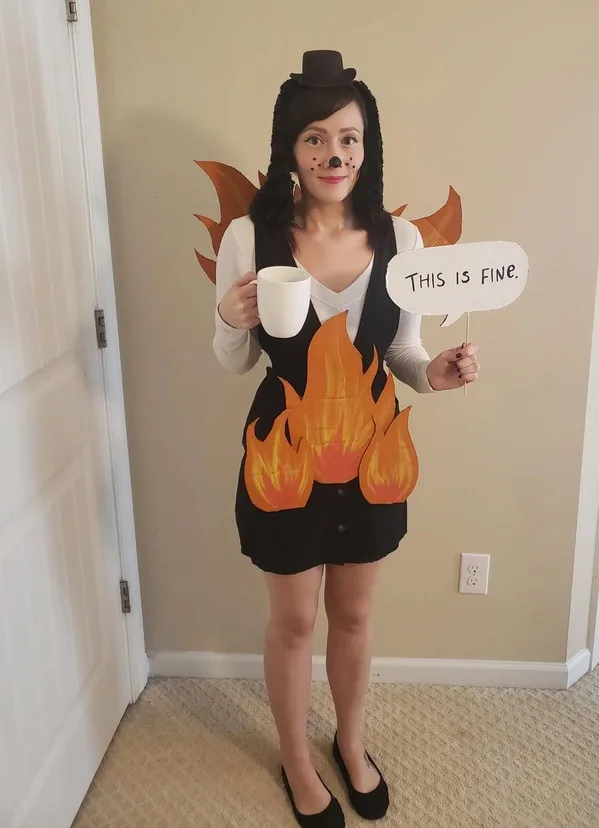 A person dressed as a fire with a black dress adorned with orange flame cutouts, wearing a top hat and holding a white mug…