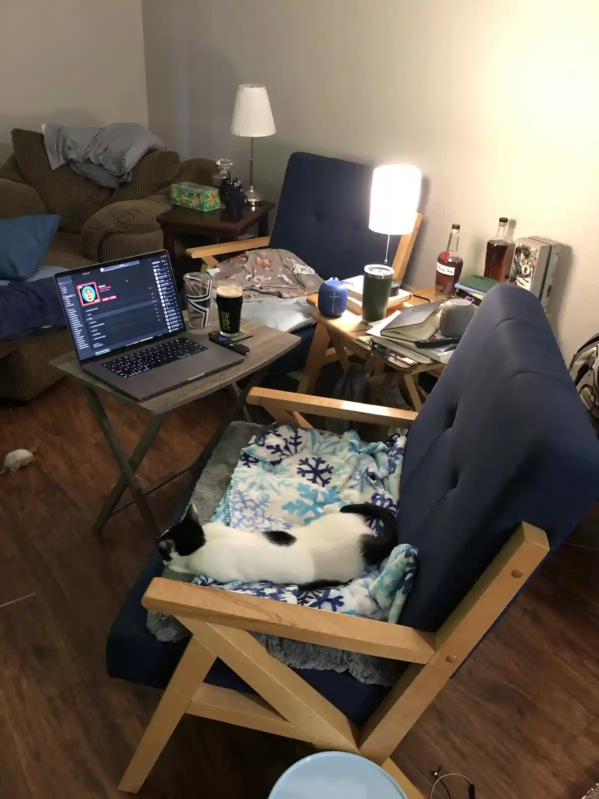 A black-and-white cat is curled up sleeping on a blue blanket in a wooden-framed recliner chair. Next to the chair is a…