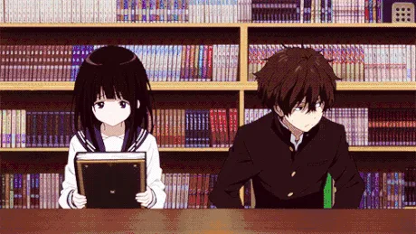 An anime scene shows a girl and boy sitting at a library table, initially both looking at their books. The girl then turns…