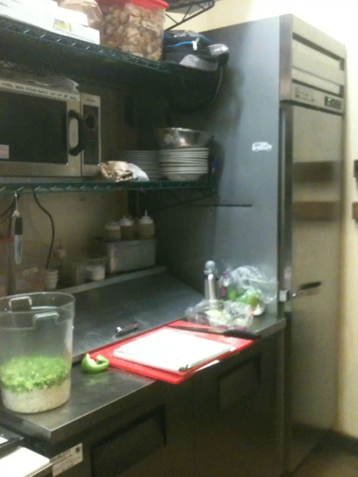 A blurry photo of a commercial kitchen with stainless steel counters, shelves holding plates and a microwave, and a large…