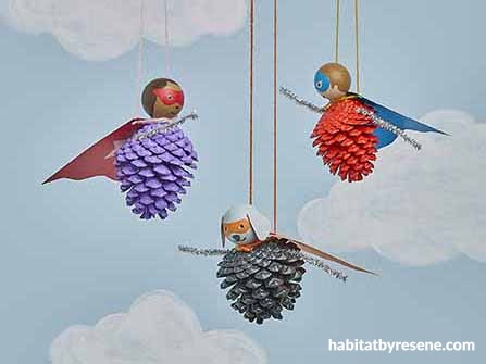 Three pinecone figures with painted faces, paper wings, and tinsel tails hang from strings against a sky with clouds. Each…
