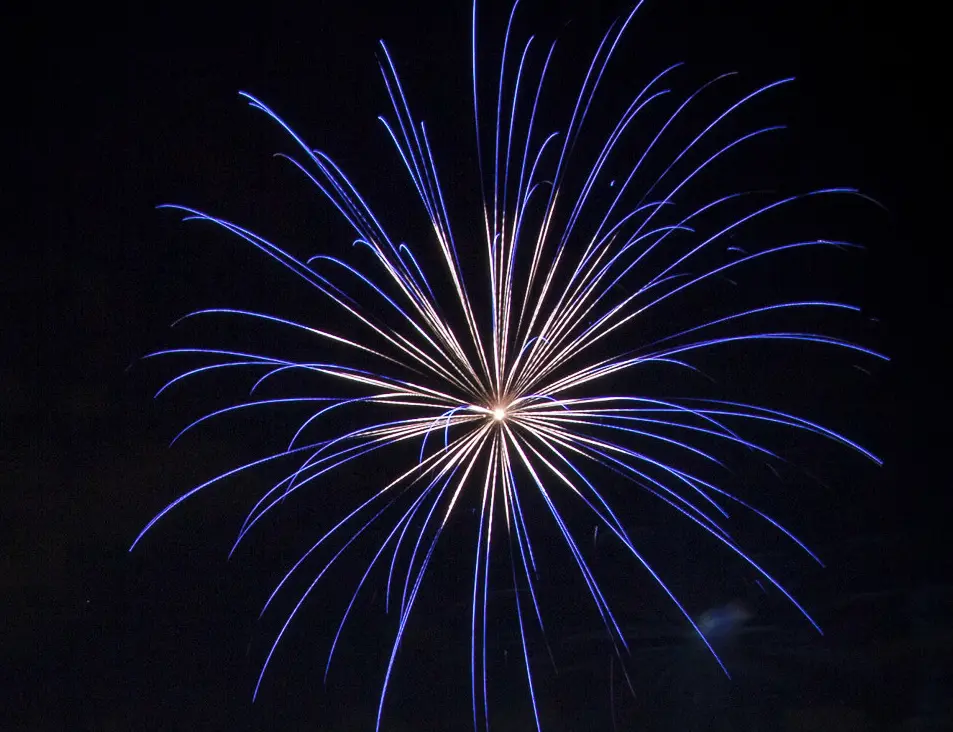 A long-exposure photograph captures a blue and white fireworks explosion against a black night sky, with streaks radiating…