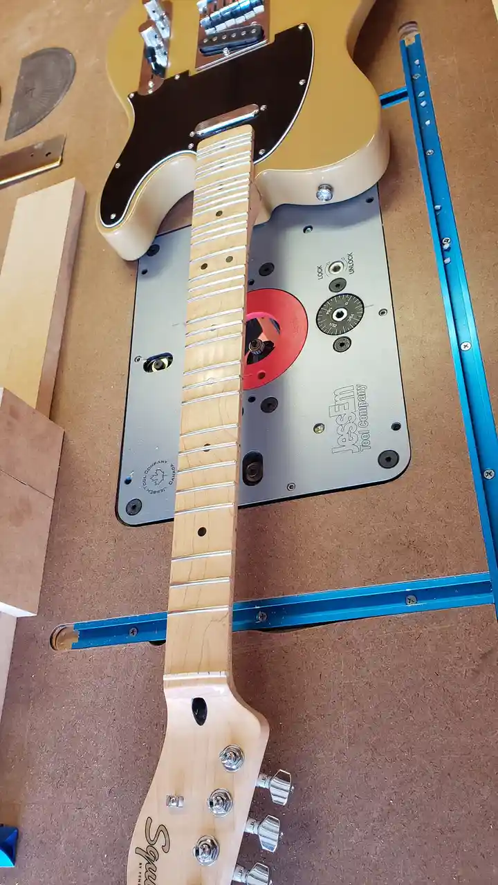 A light-colored electric guitar body and neck are positioned over a Jescem router table, with the neck aligned over the…