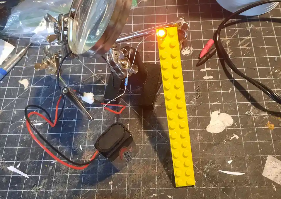 A yellow Lego brick stands upright on a gridded workbench, with a small LED light glowing at its top. Wires connect the LED…