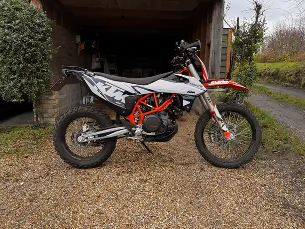 A white, orange, and black KTM Enduro R dirt bike is parked on gravel in front of a wooden garage. The bike has prominent…