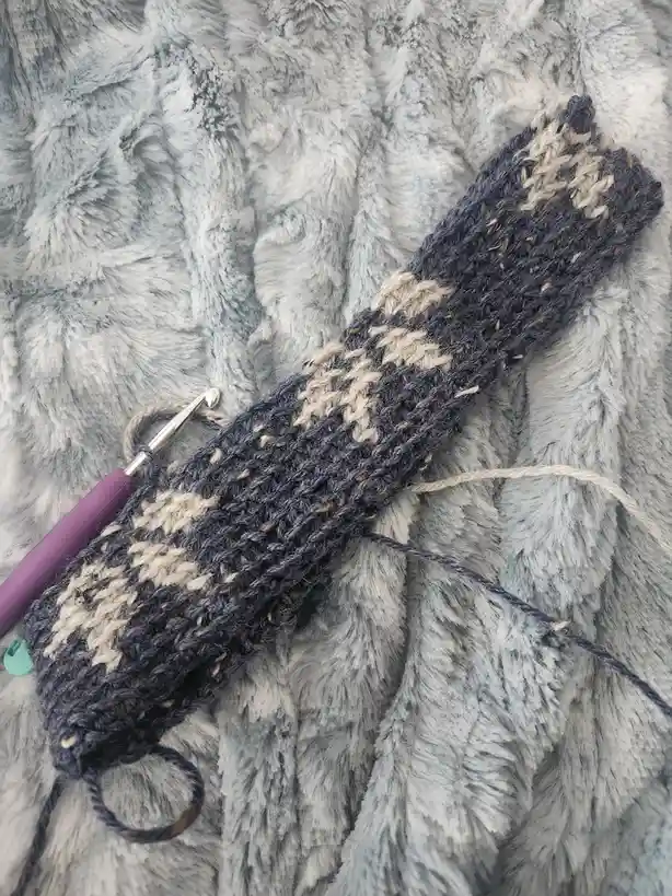 A partially completed crocheted item, likely a scarf or hat, in dark blue and cream yarn, lies on a fluffy gray blanket with…