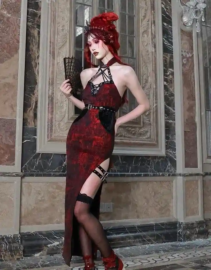 A woman with red hair and gothic makeup poses in a red and black lace dress with thigh-high stockings and a black fan…
