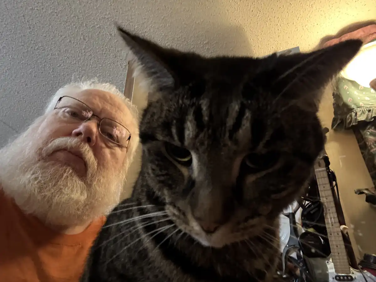 A selfie shows an older man with a white beard and glasses next to a large tabby cat, both looking at the camera. The cat…