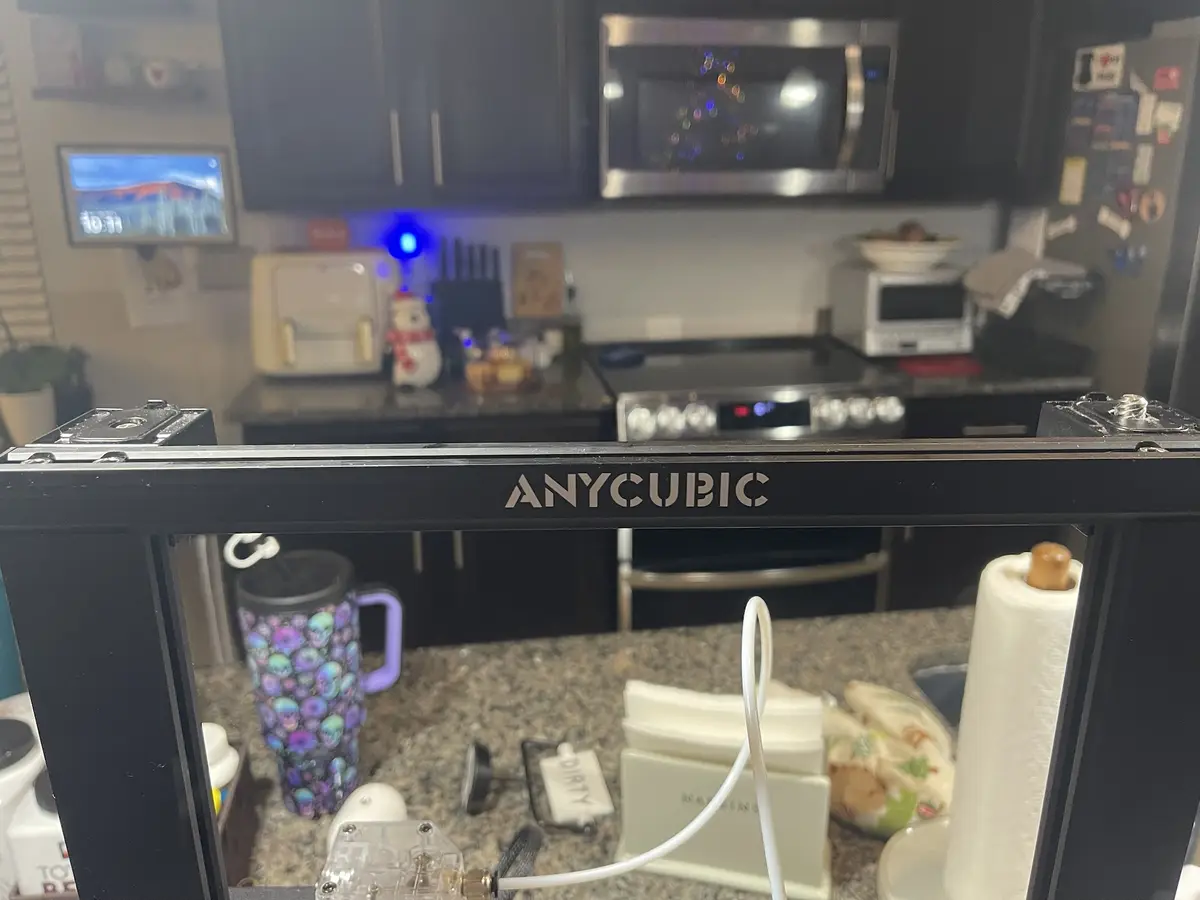 A close-up photo shows the front of a black Anycubic 3D printer sitting on a kitchen countertop, with a blurry kitchen…