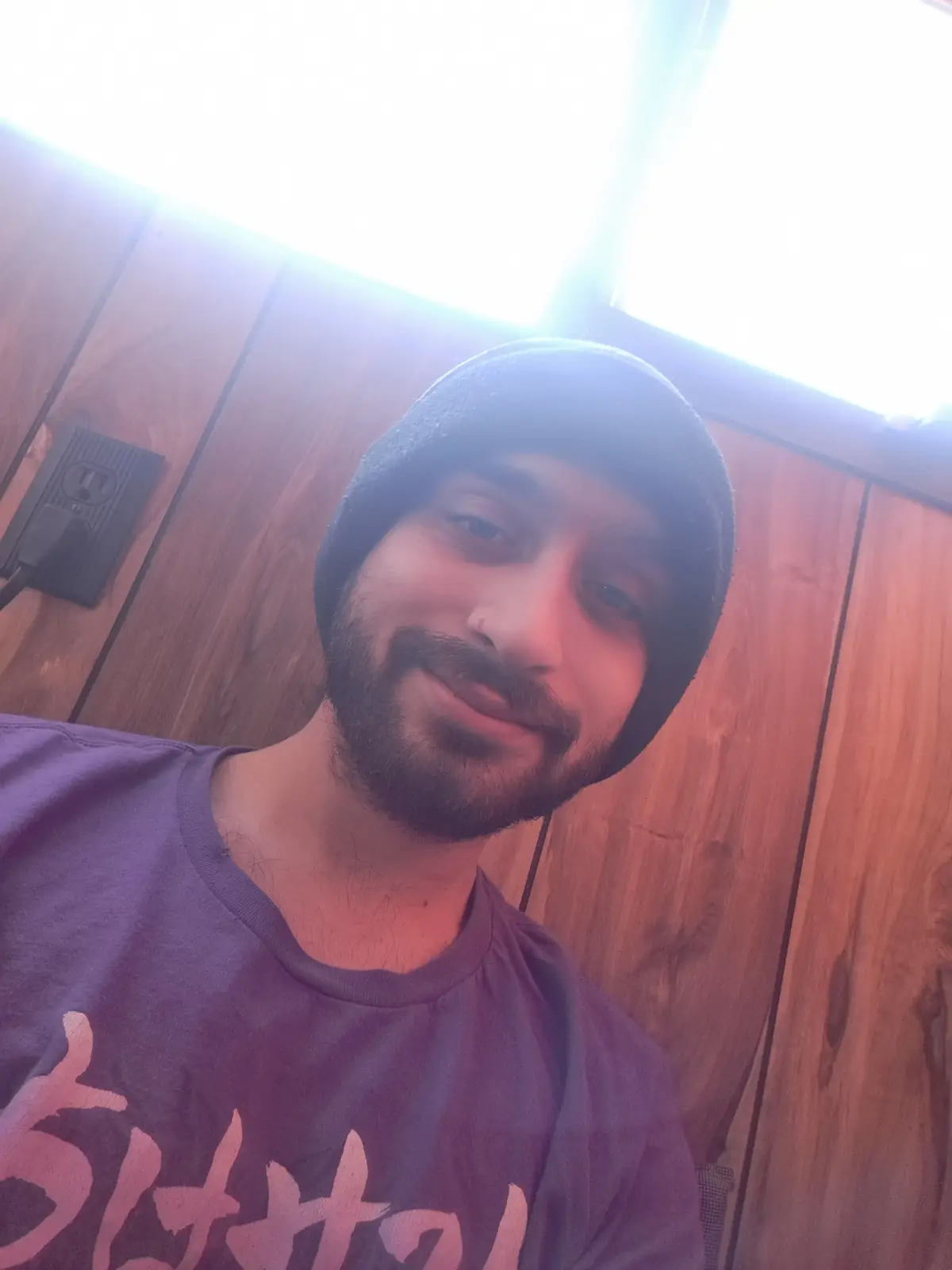 A selfie of a man with a beard and beanie, smiling slightly while looking at the camera, taken indoors against a wooden wall…