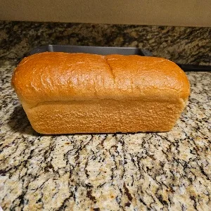 A golden-brown, rectangular loaf of bread sits on a speckled granite countertop, with a metal loaf pan visible behind it…