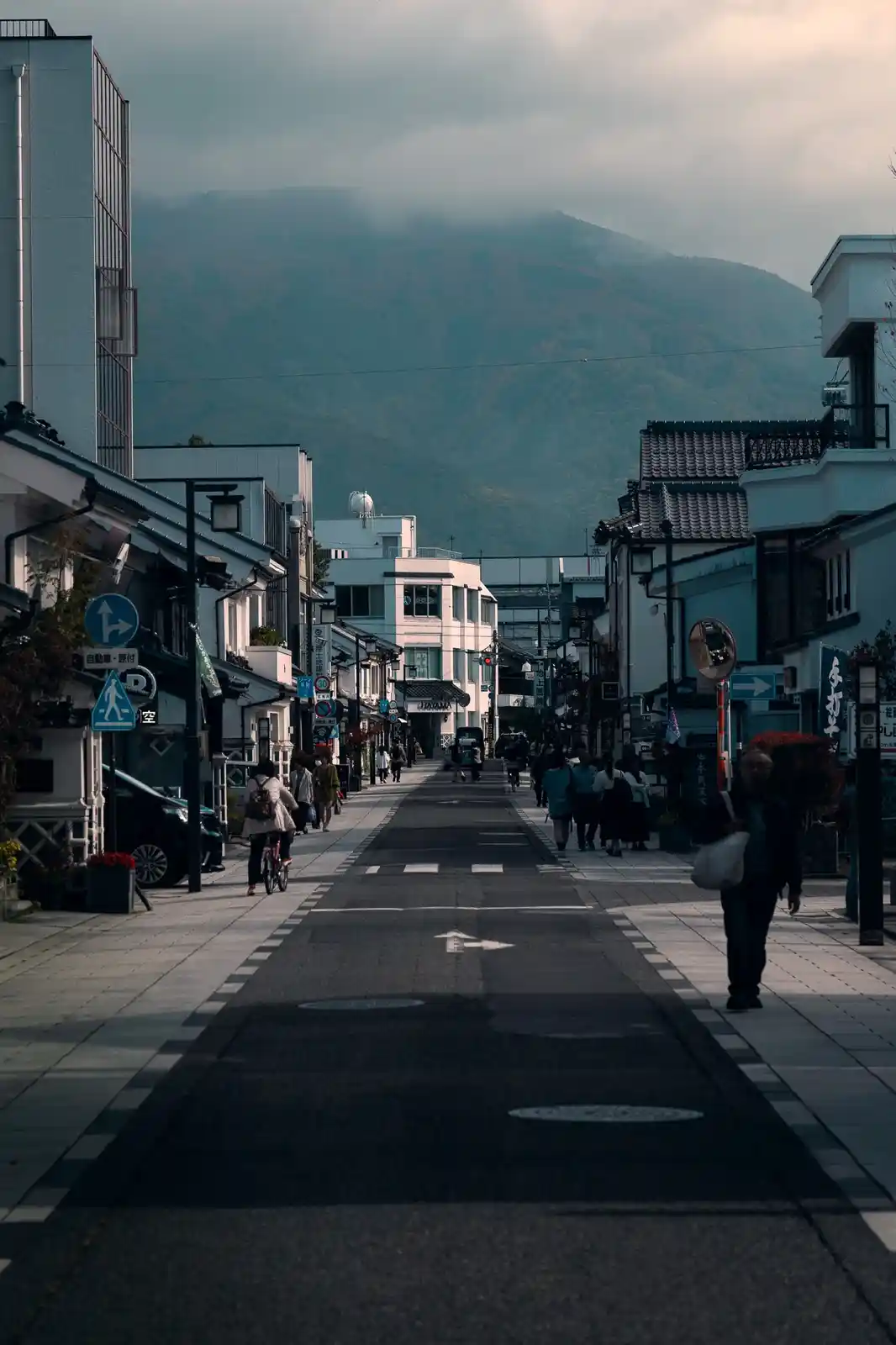 A serene, narrow street in a Japanese town with traditional and modern buildings, pedestrians walking, and a mountain…