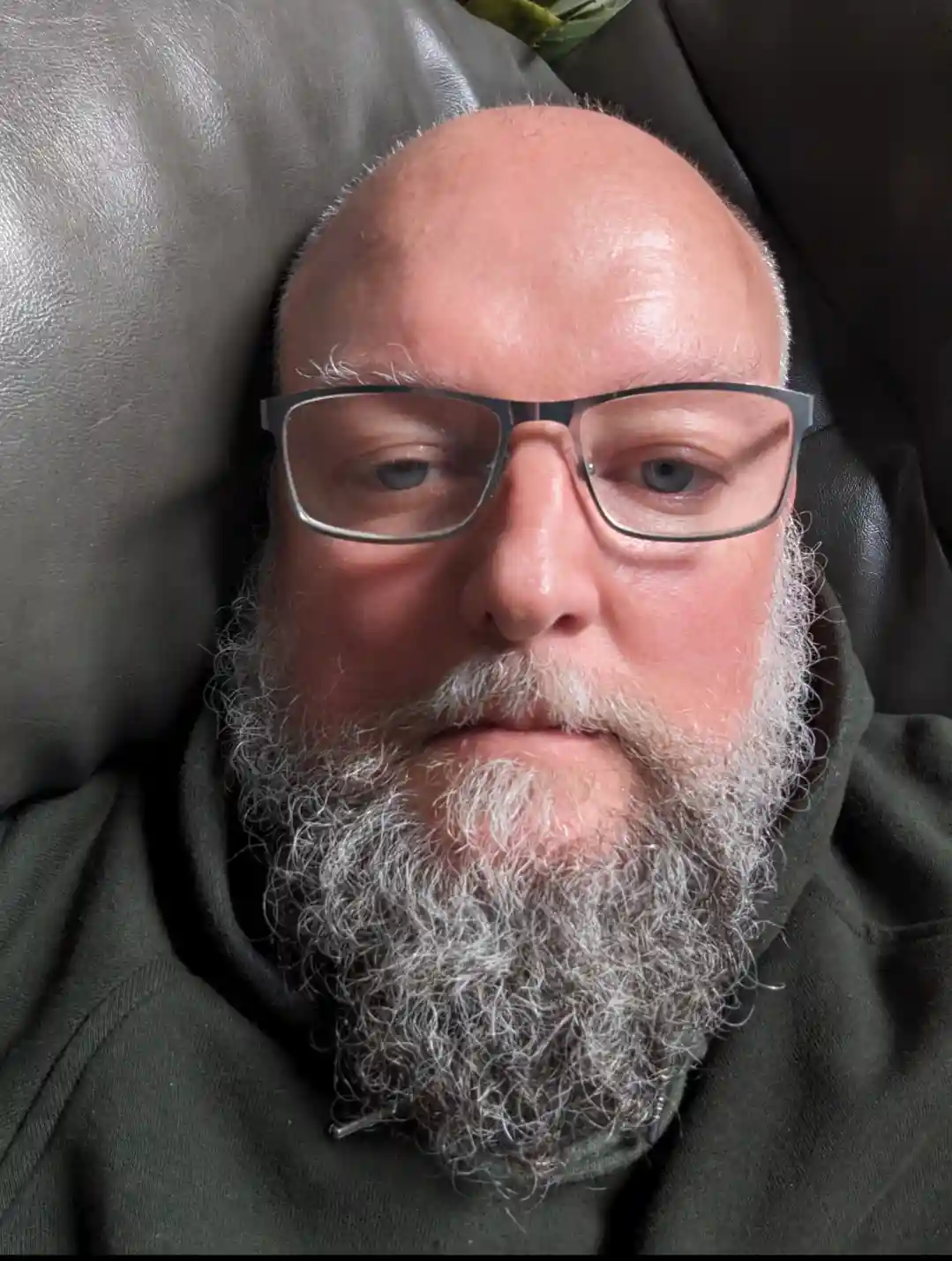 A close-up photo of a middle-aged man with a bald head, thick gray beard, and glasses, lying back on a dark leather couch…