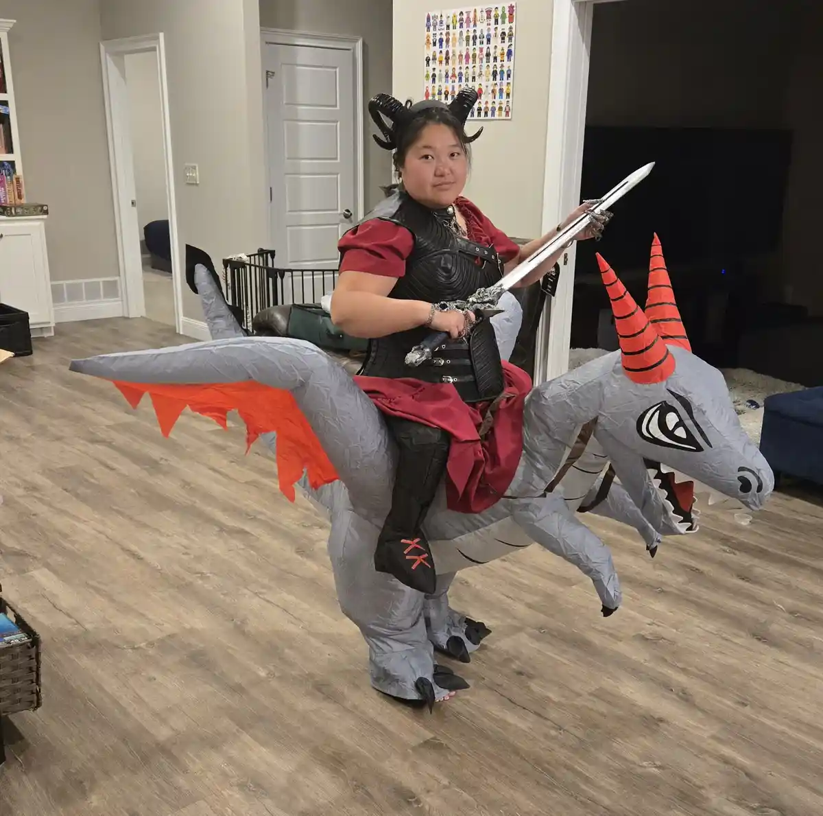 A person wearing a red dress, black armor, and black horns sits atop a large, gray inflatable dragon costume with orange…
