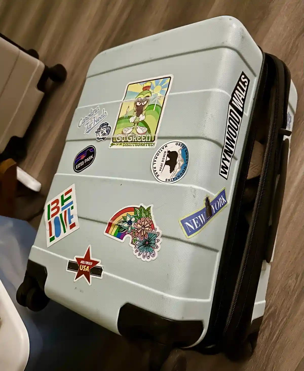 A light blue hard-shell suitcase covered in numerous travel stickers, including 'Wynwood Walls', 'New York', 'Hollywood…