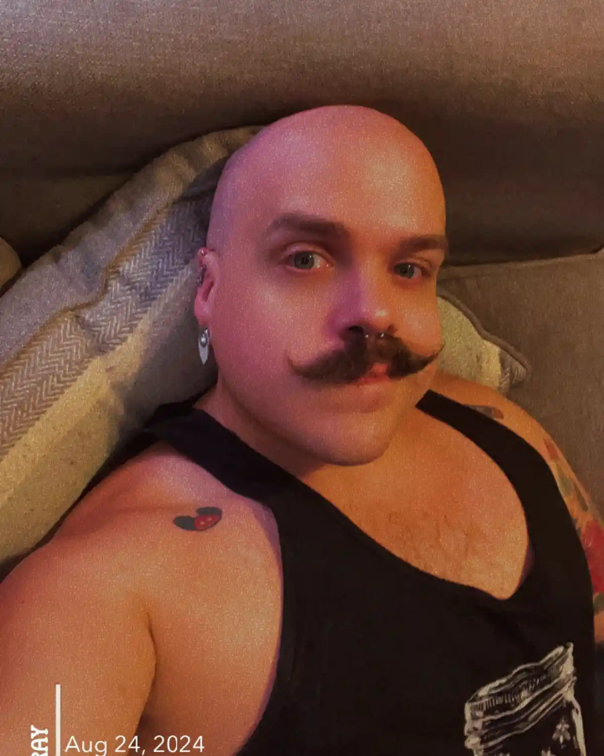 A bald man with a thick mustache, nose ring, and ear piercings lies on a couch wearing a black tank top. A small ladybug…