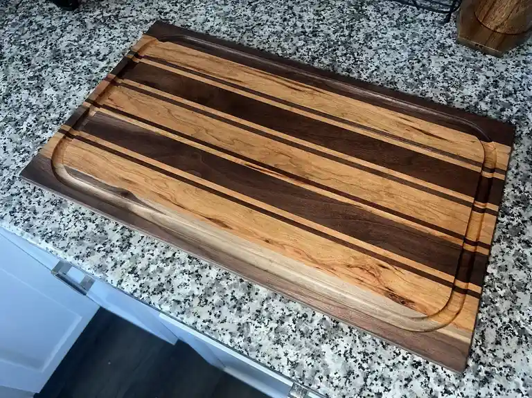 A high-quality, striped wooden cutting board with alternating light and dark wood planks rests on a speckled granite kitchen…
