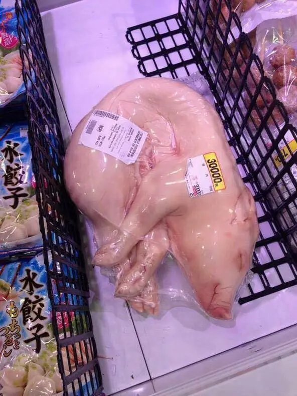 A whole, raw, packaged pig is displayed in a black plastic grocery basket in a refrigerated section, with a yellow price tag…