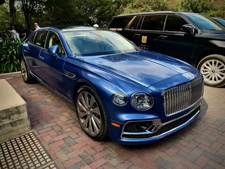 A shiny blue Bentley sedan is parked on a brick driveway, with a black limousine behind it that has 'HOTEL EMMA' and a crest…
