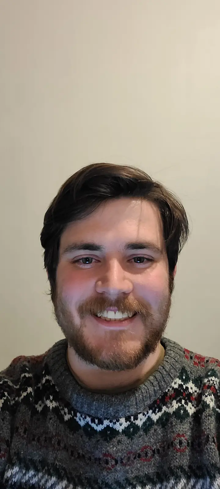 A man with brown hair, a beard, and mustache smiles while wearing a patterned knit sweater against a plain wall. The…
