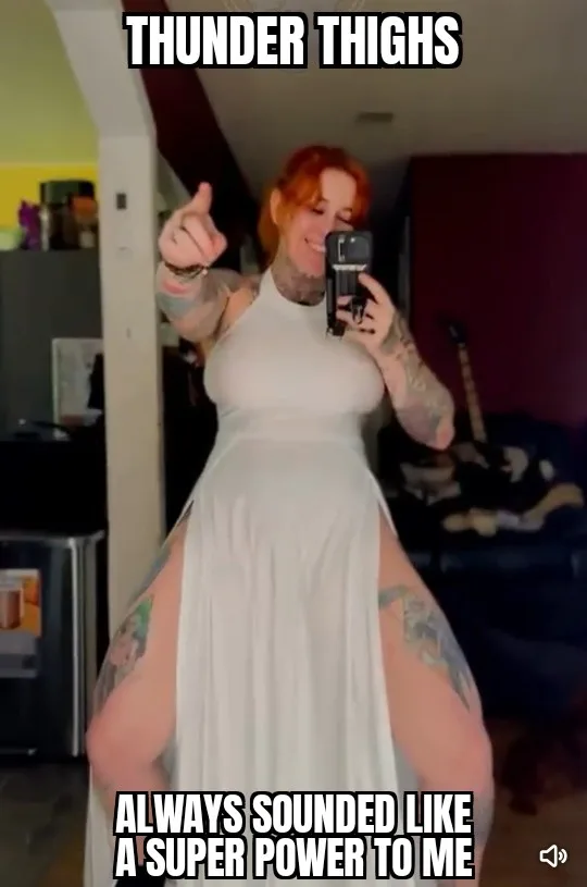A woman with red hair and tattoos takes a mirror selfie in a white dress with high slits, pointing at the camera. The text…