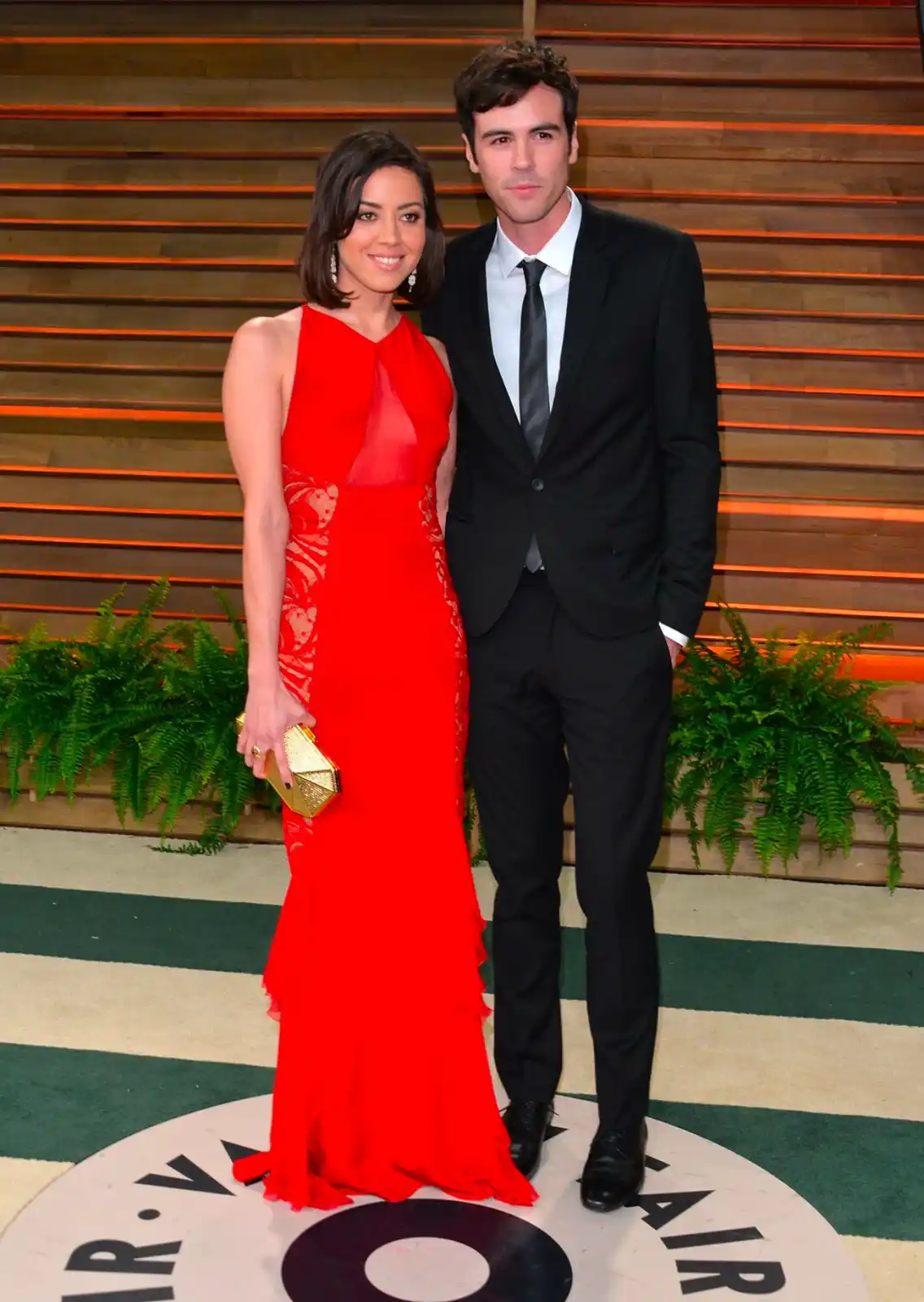 A man and woman pose together at a red carpet event, the woman in a striking red dress and the man in a black suit. The…