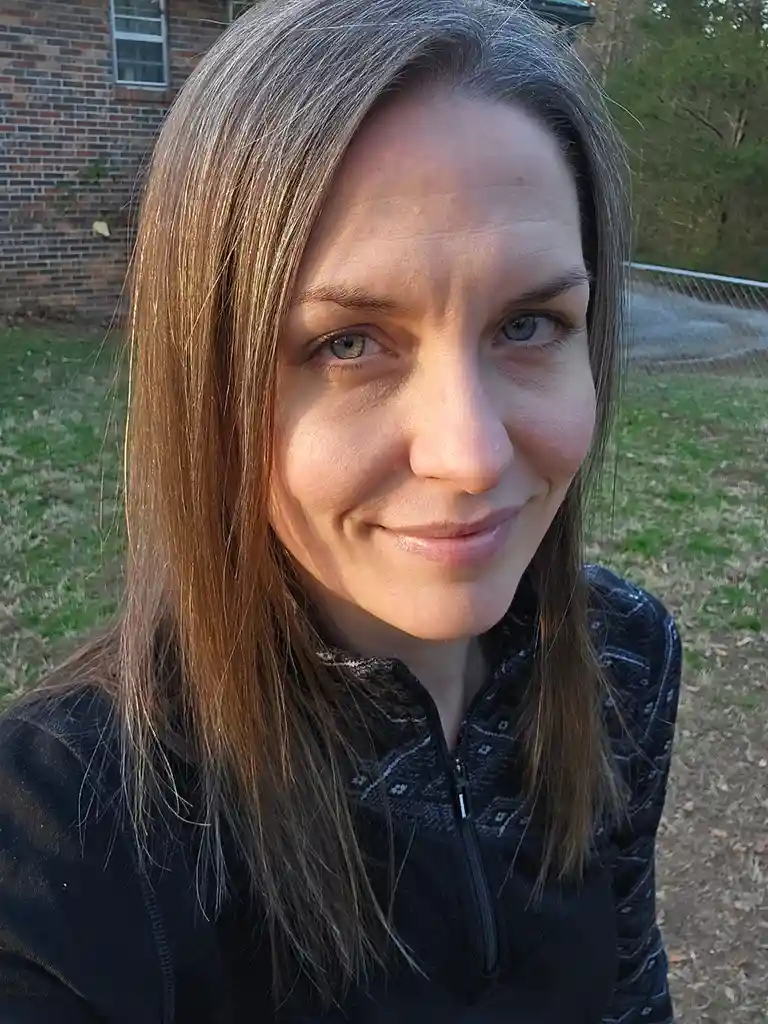 A woman with brown hair and gray streaks smiles at the camera while wearing a black zip-up jacket outdoors. Behind her is a…