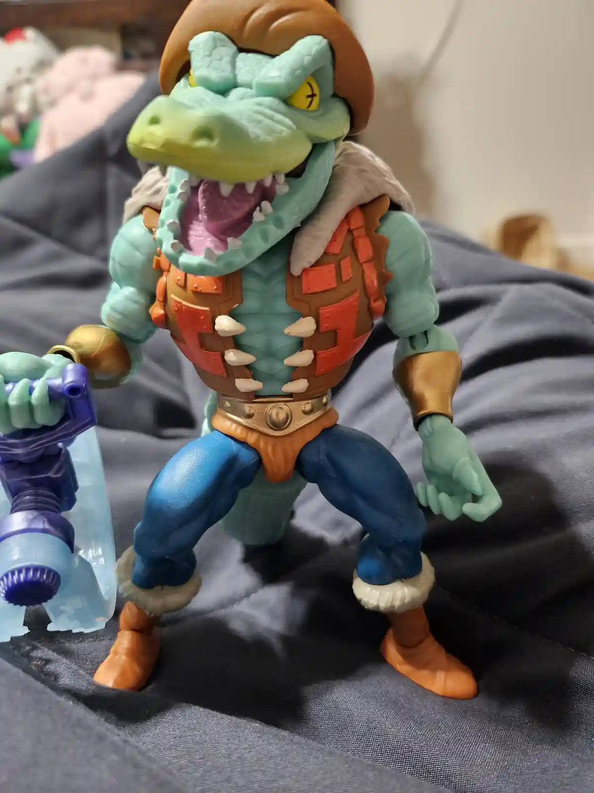 A close-up photo of a green crocodile-like action figure with a brown hat, orange and brown armor, and blue pants, standing…