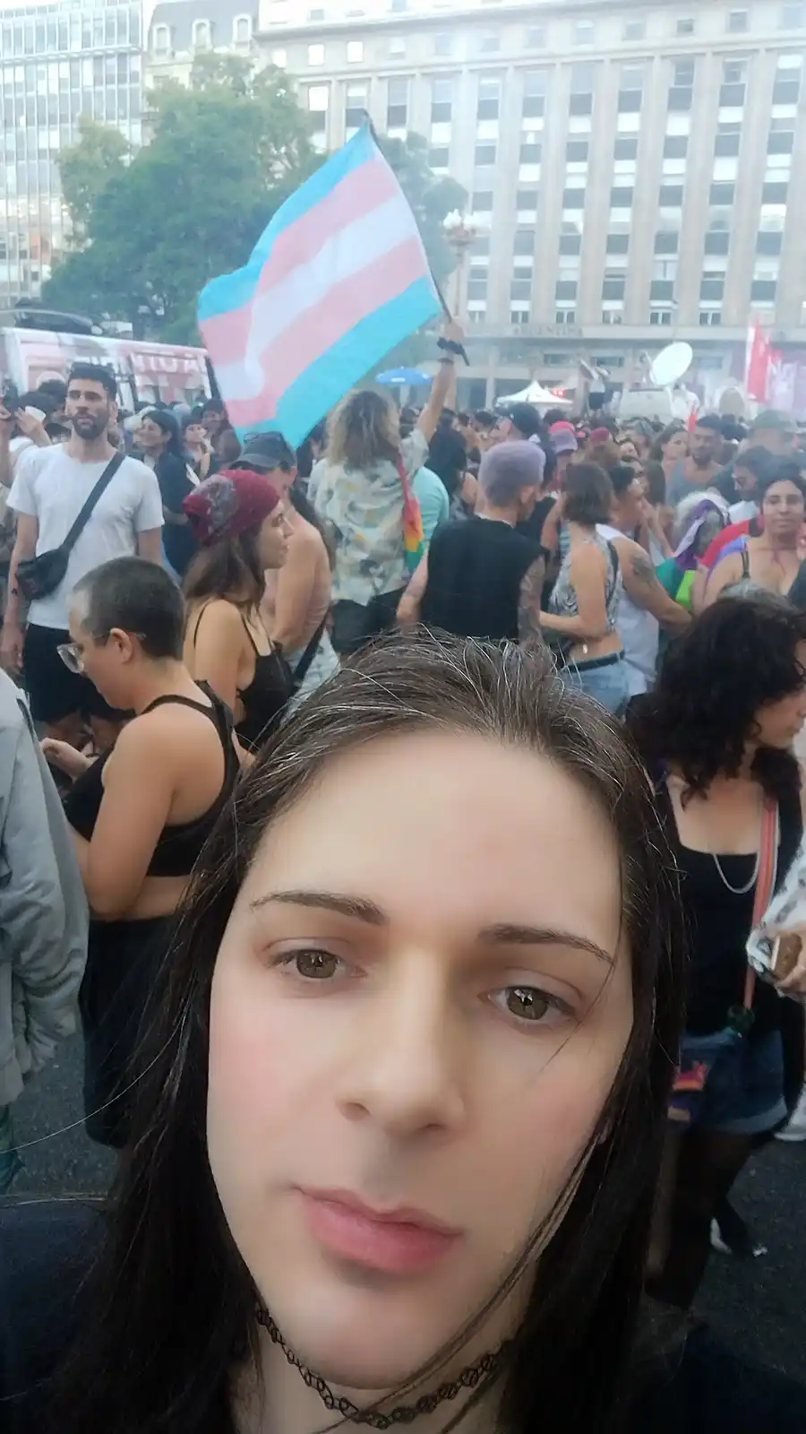 A selfie of a person with long dark hair and a choker, looking directly at the camera with a neutral expression, taken at a…