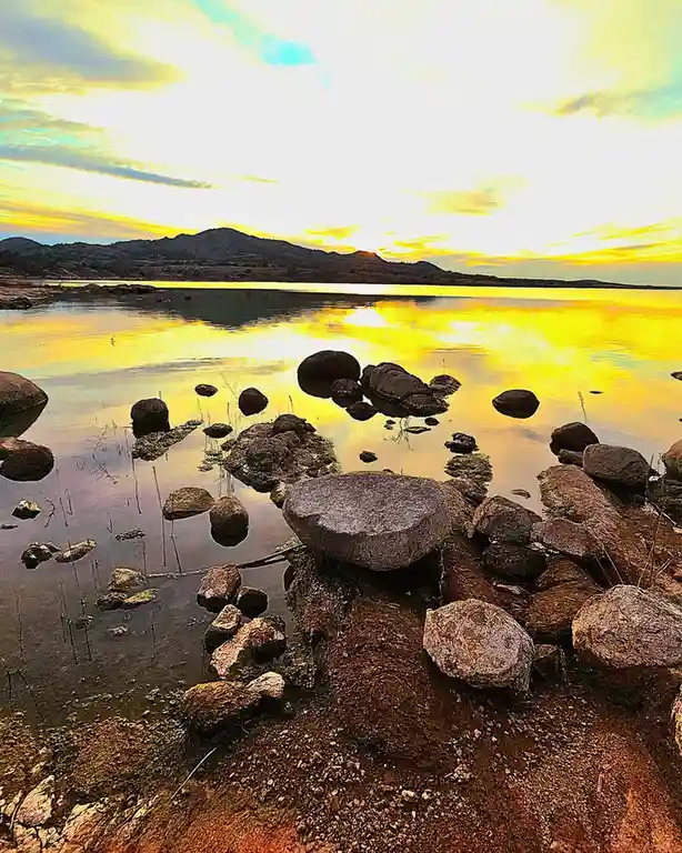 A serene landscape photo of a lake at sunset with rocks in shallow water, reflecting golden light, overlaid with the text…