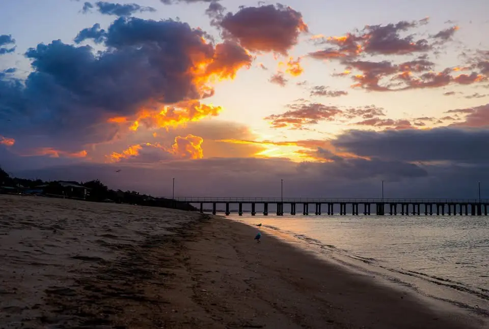 A serene beach sunset with vibrant orange and purple clouds, a long pier extending into the water, and a lone person in a…