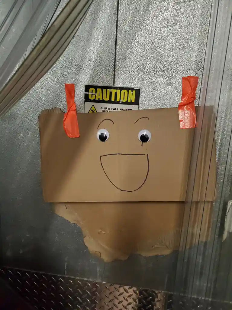 A brown cardboard box with googly eyes and a hand-drawn smiley face is taped to a glass door with orange duct tape. Behind…