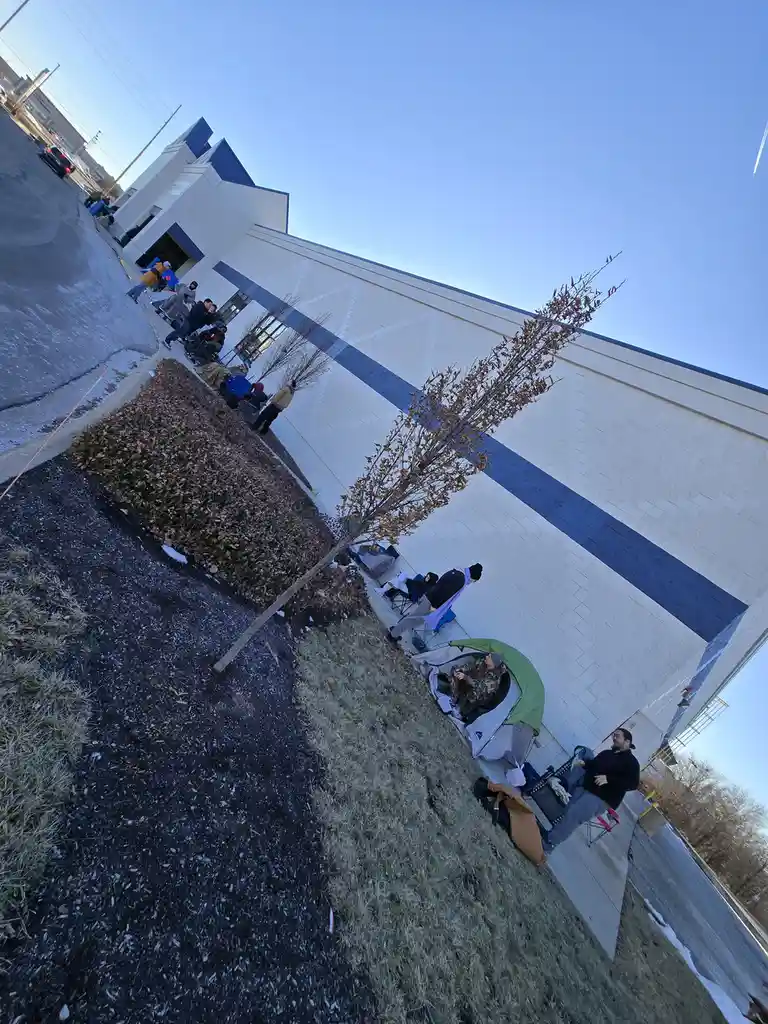 A tilted photo shows people camping outside a white building with blue stripes, some in chairs, one in a tent, on a sidewalk…