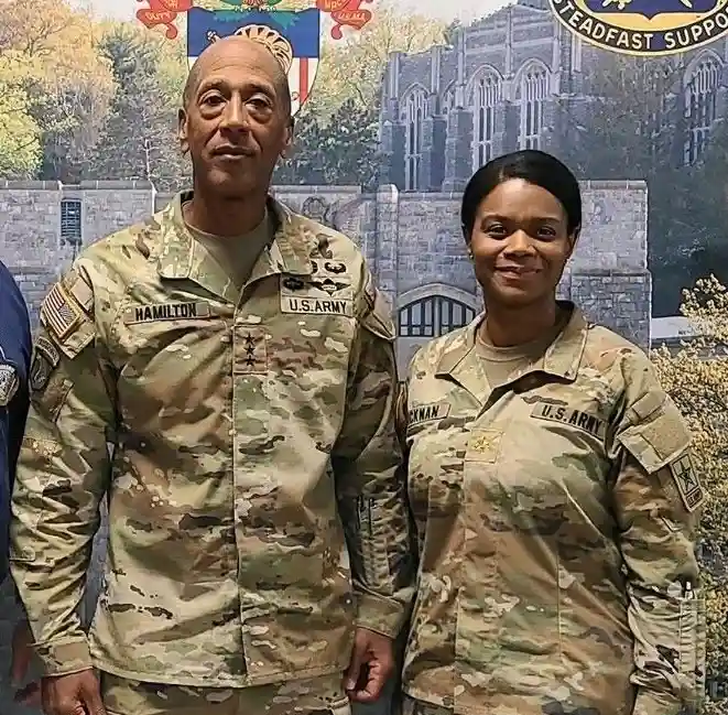 Two U.S. Army officers in camouflage uniforms stand side by side in front of a backdrop featuring a castle and military…