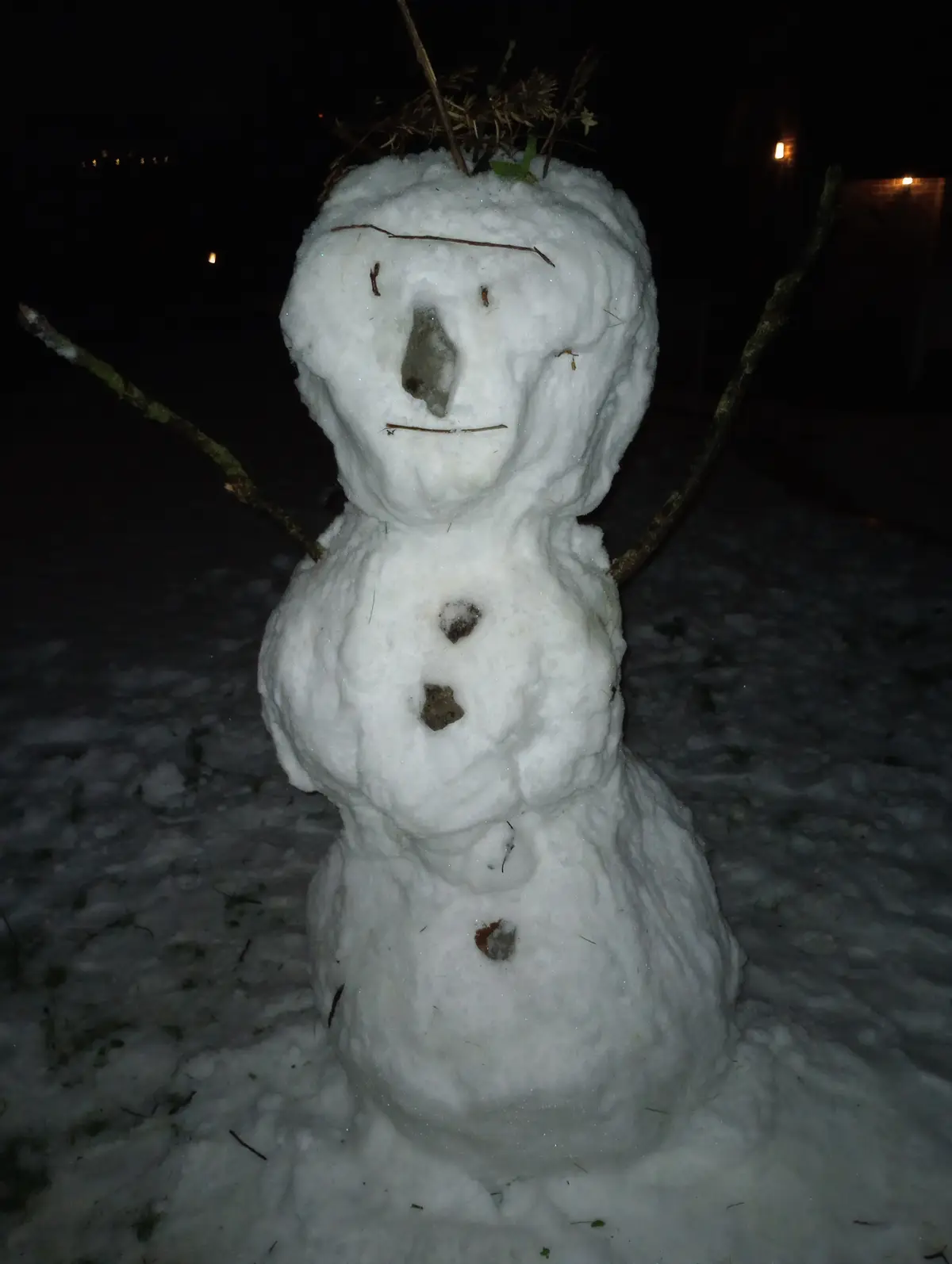 A snowman built at night with a face made of twigs and a rock for a nose, topped with dried grass, standing in snow with…