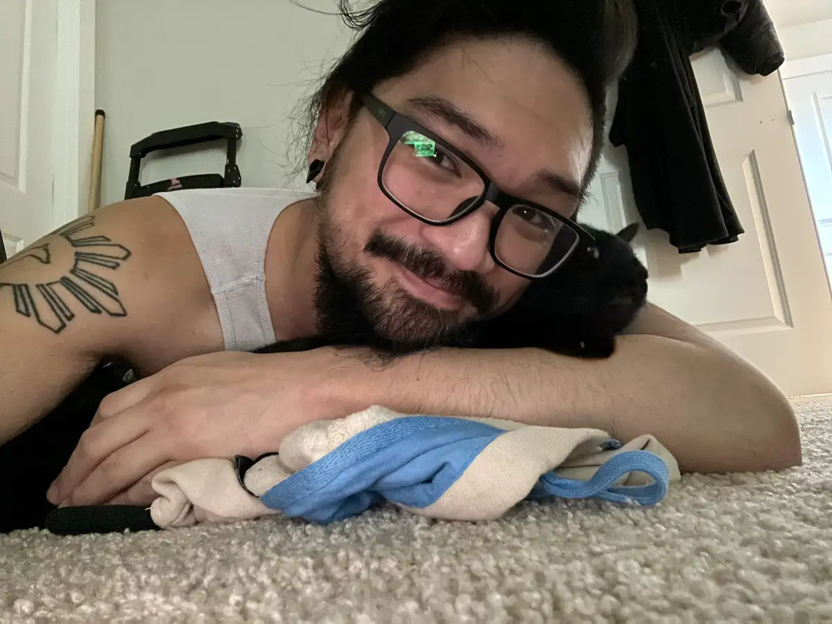 A man with glasses and a sun tattoo on his arm lies on a carpeted floor, smiling while holding a black cat. A blue and white…