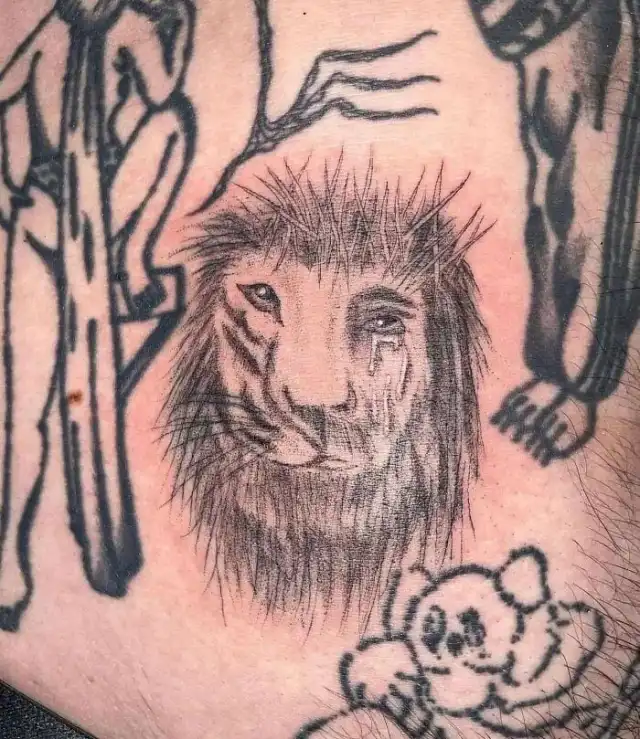 A close-up of a tattoo on human skin showing a detailed, sorrowful lion's face with a tear rolling down its cheek…