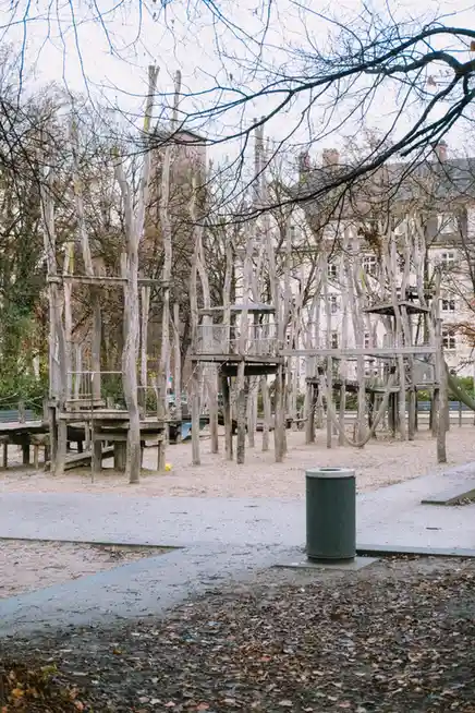 a photograph of an empty, leafless playground with bare trees and wooden structures, viewed through bare branches, with a…