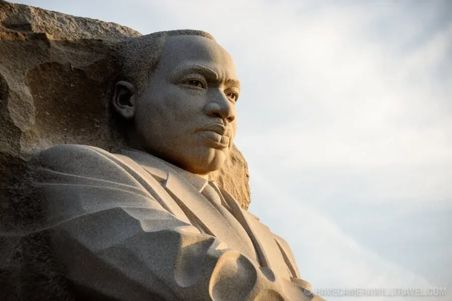 A close-up photograph of the stone sculpture of Martin Luther King Jr., showing his face and shoulders against a cloudy sky…