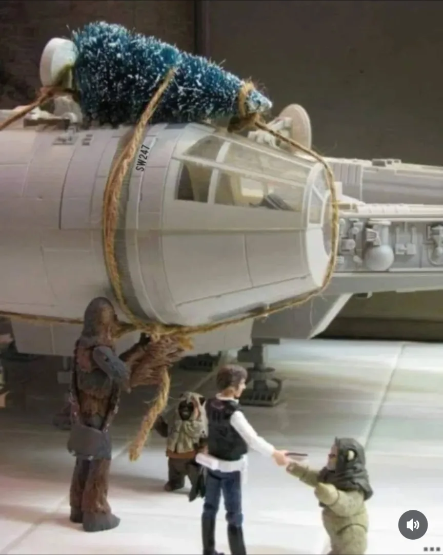 A toy model of the Millennium Falcon has a small blue tinsel Christmas tree strapped to its top with twine. Below, toy…