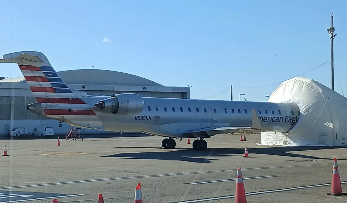 An American Eagle regional jet, registration N585NN, is parked on an airport tarmac with its nose covered by a large white…