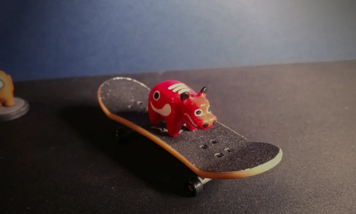 A small red demon figurine with white markings and black horns is positioned on a miniature skateboard with black grip tape…