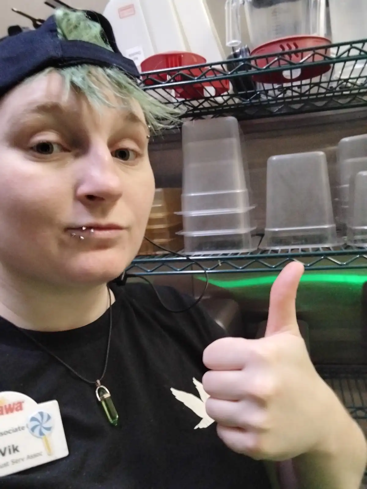 A person with green-tinted hair and a black cap gives a thumbs-up while wearing a name tag that reads 'Vik, Just Serv Assoc'…