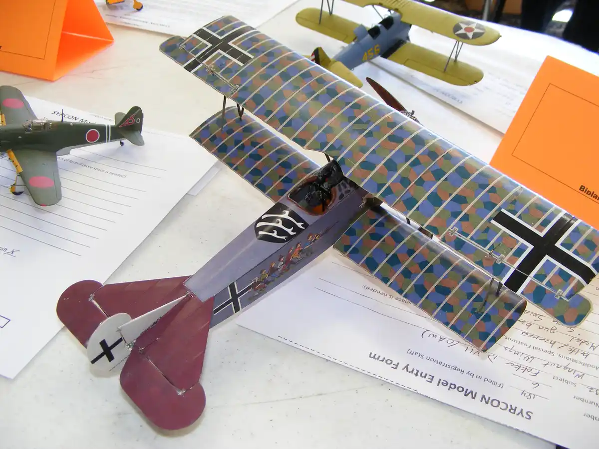 A model biplane with German military markings and a cartoonish decal of children on its fuselage sits on a 'SYRCON Model…