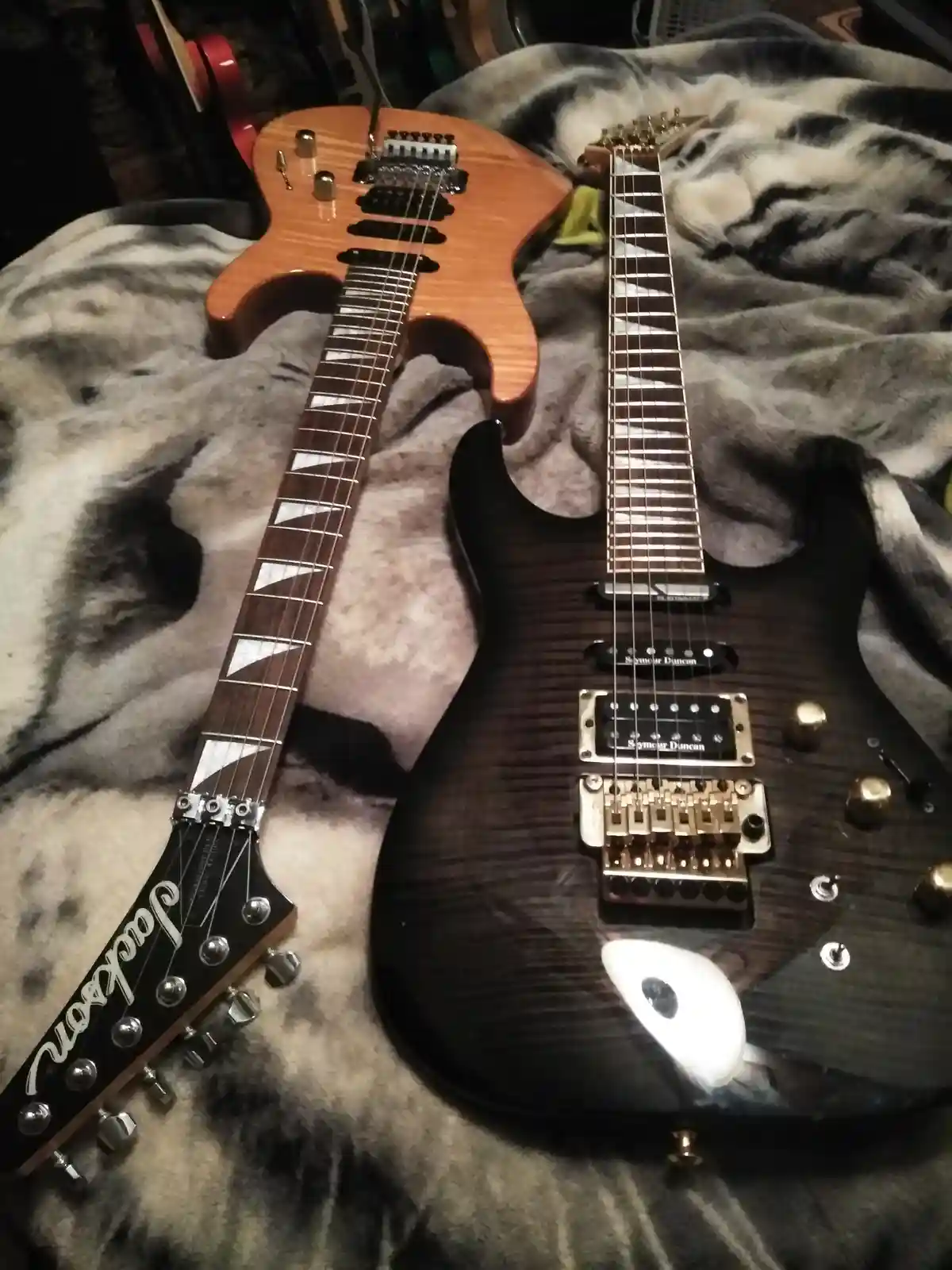 Two electric guitars lie side by side on a gray and black patterned blanket; one is a light wood-finish Jackson with a…