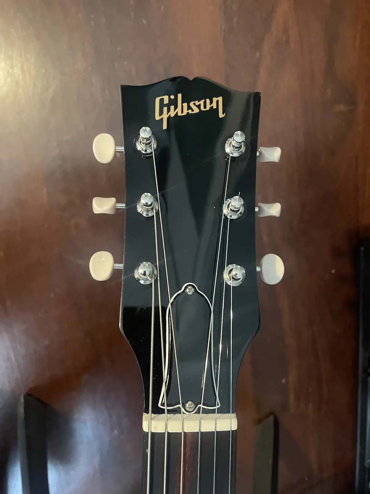 A close-up photo of a Gibson guitar headstock with six tuning pegs and strings, set against a dark wood background. The…
