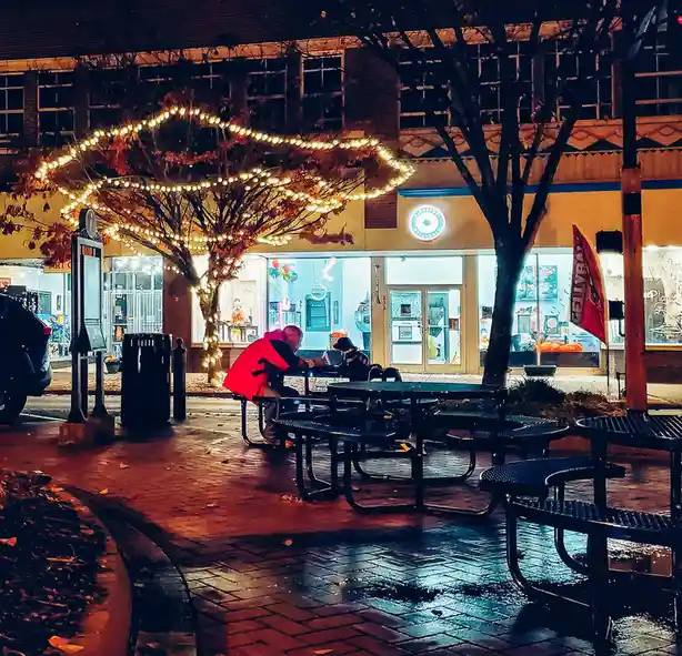 A nighttime photo of two people sitting at an outdoor picnic table in front of a brightly lit storefront. One person wears a…