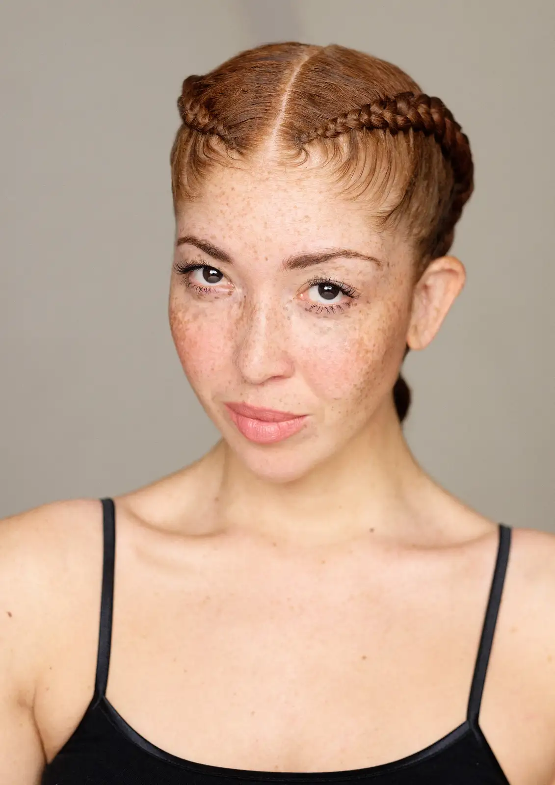 A woman with freckles and reddish-brown hair styled in two braids along her part looks directly at the camera with a neutral…