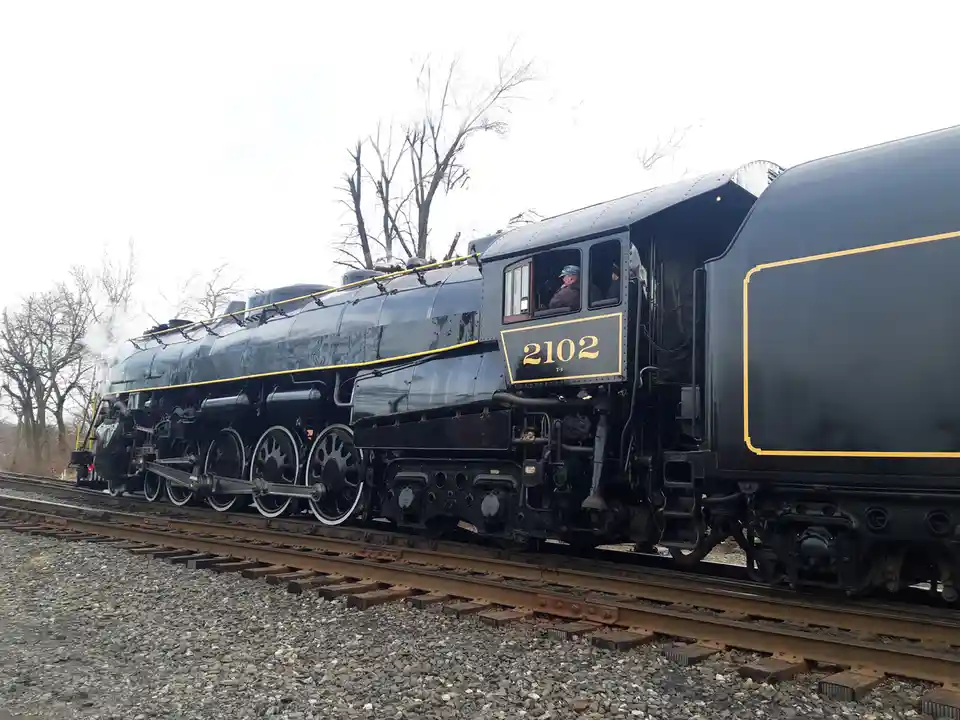 A black steam locomotive with yellow trim and the number 2102 on its side is shown on railway tracks, with bare trees in the…